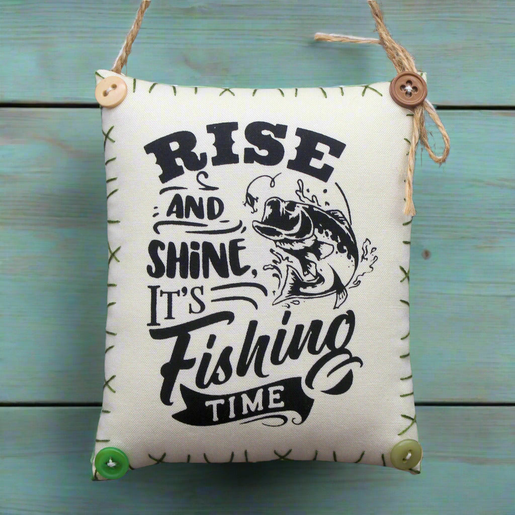 Hanging mini pillow with 'Rise and Shine, It's Fishing Time' text and fish illustration on a green wooden background