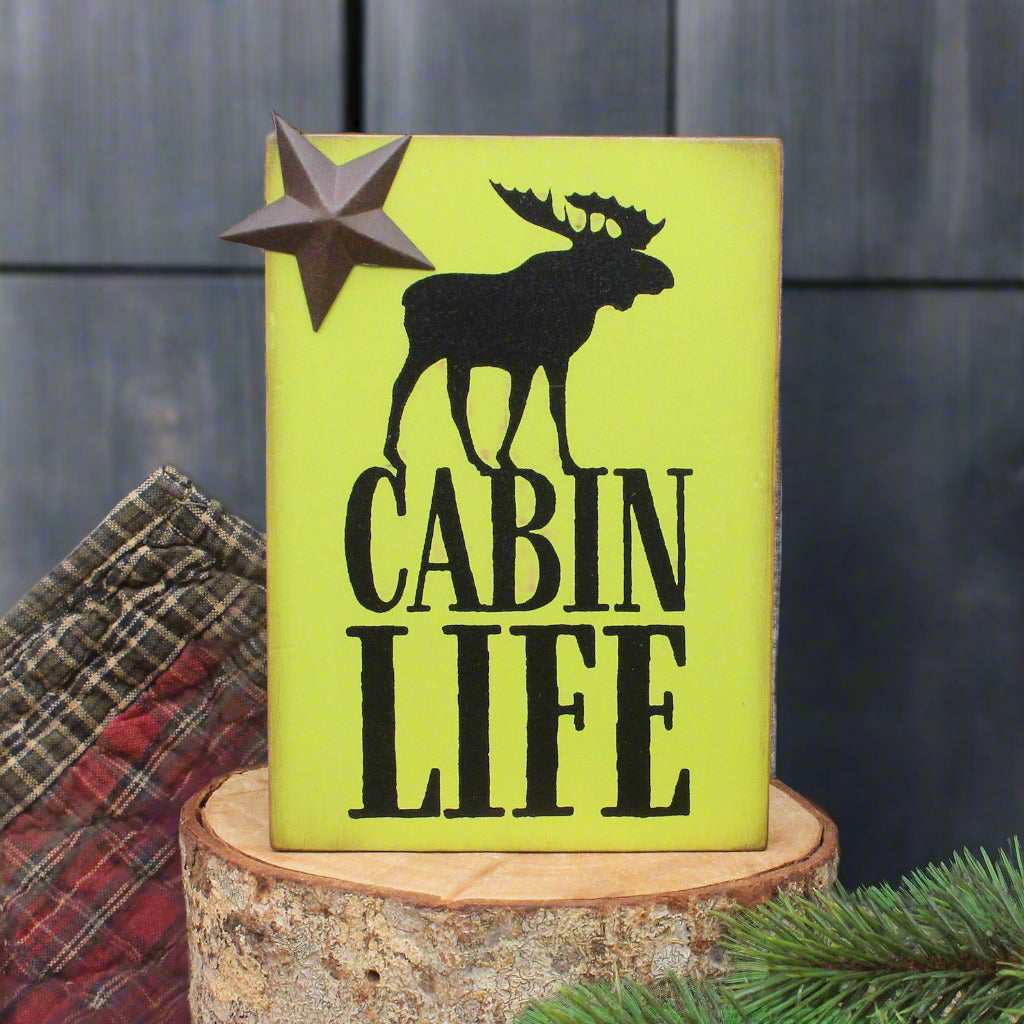 Wooden sign with moose silhouette and 'Cabin Life' text on a wooden stump with plaid fabric and pine branches.