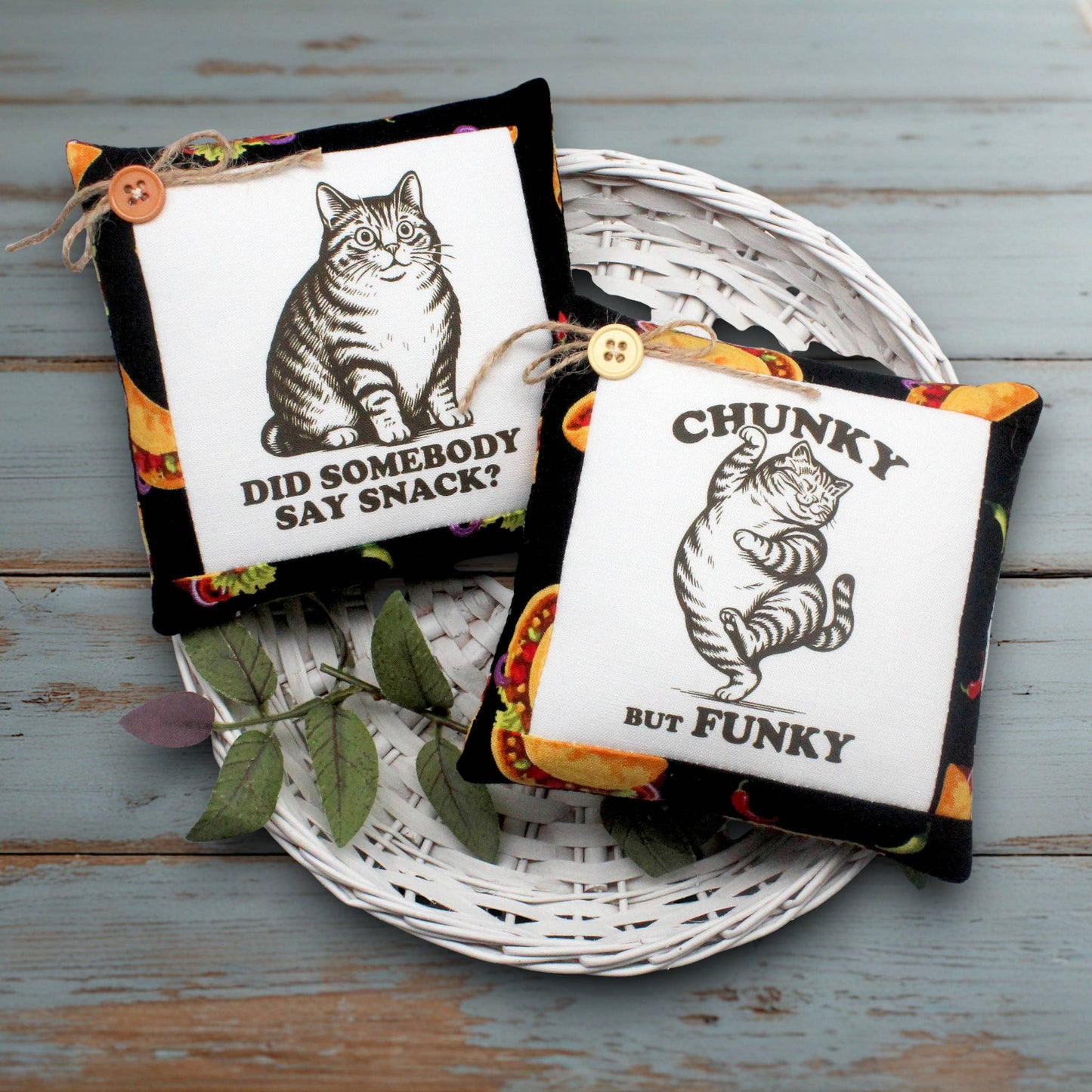 Two decorative square fabric mini pillows displayed on a white wicker tray over a distressed blue-gray wooden surface. Each pillow has a black border, a white center panel, a small wooden button, and a colorful patterned edge.