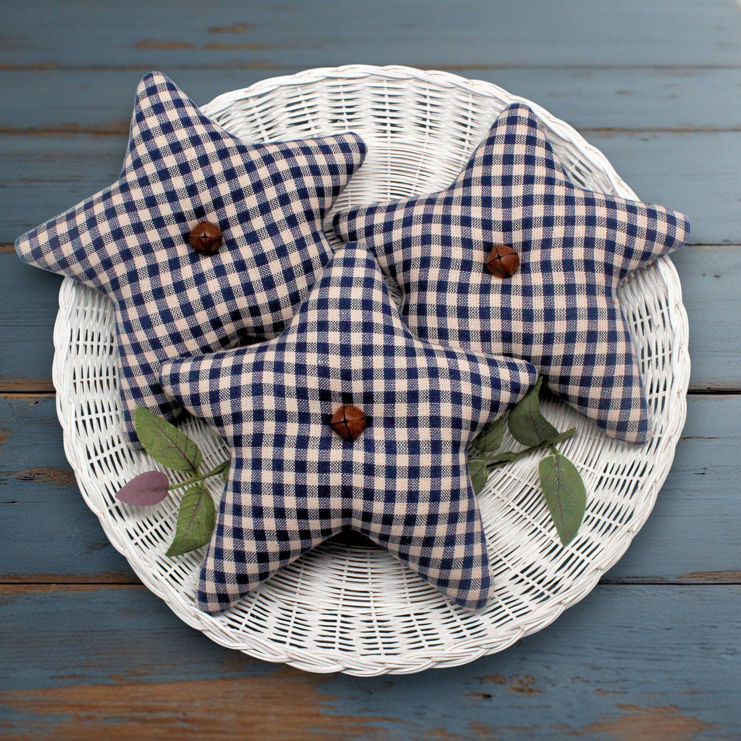 Blue and beige checkered fabric star bowl fillers with rusty bells on a white wicker basket against a blue wooden background.