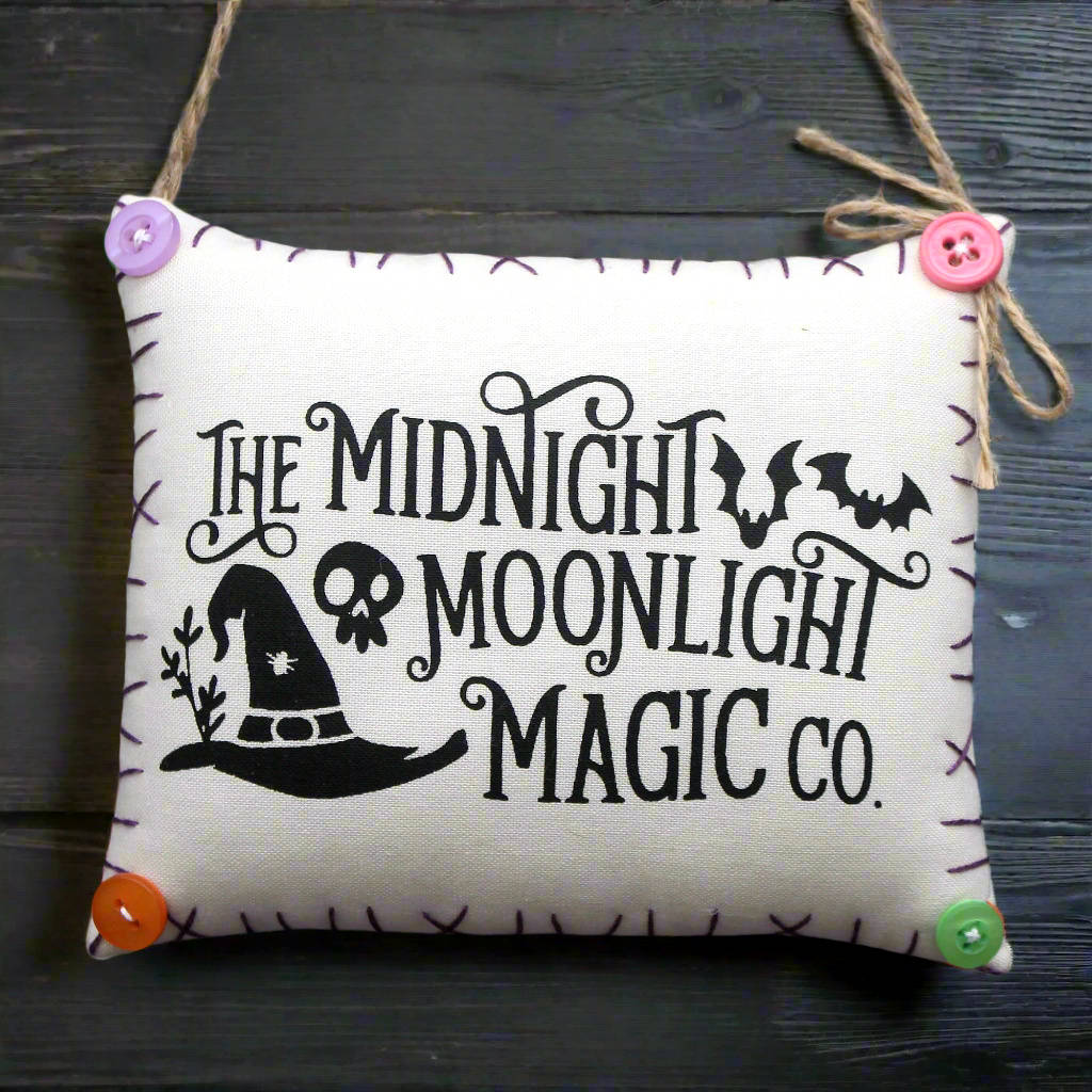 Decorative pillow with 'The Midnight Moonlight Magic Co.' text and witch hat design on a wooden surface.