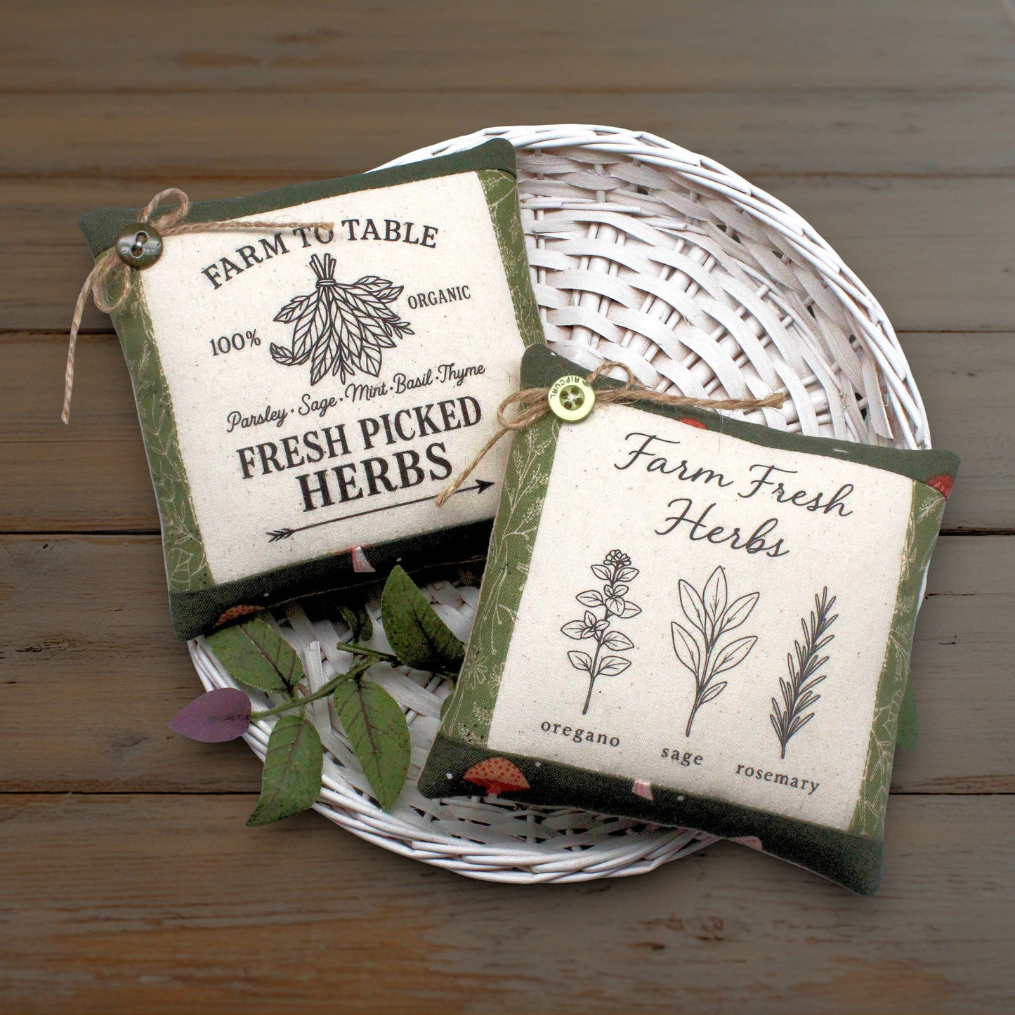 Two small fabric herb-themed mini pillows displayed on a white woven tray over a rustic wooden surface. One reads 'Farm to Table' with an illustration of a herb bunch, and the other reads 'Farm Fresh Herbs' with illustrations of oregano, sage, and rosemary.