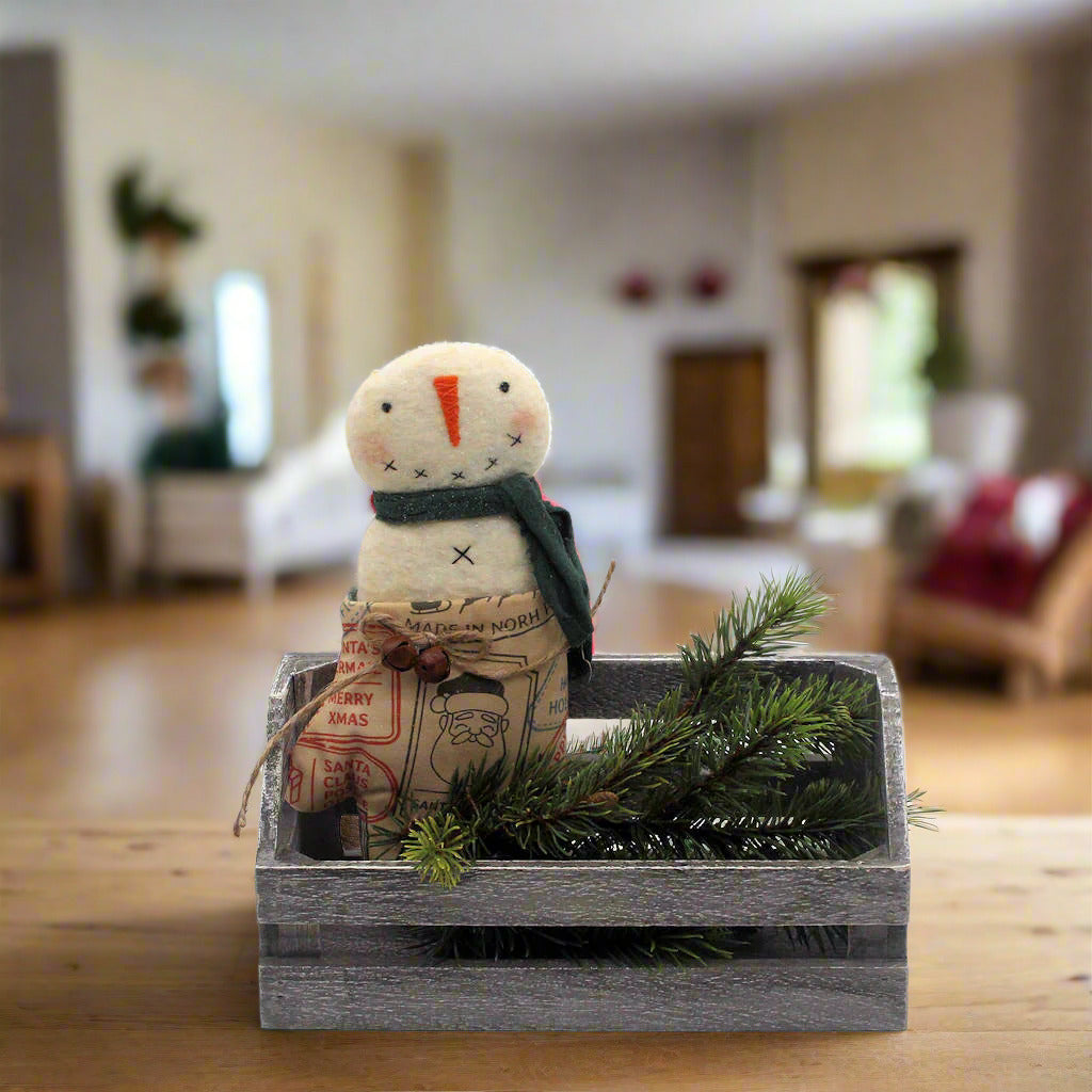 Small snowman figurine with a scarf and newspaper, sitting in a wooden crate with greenery