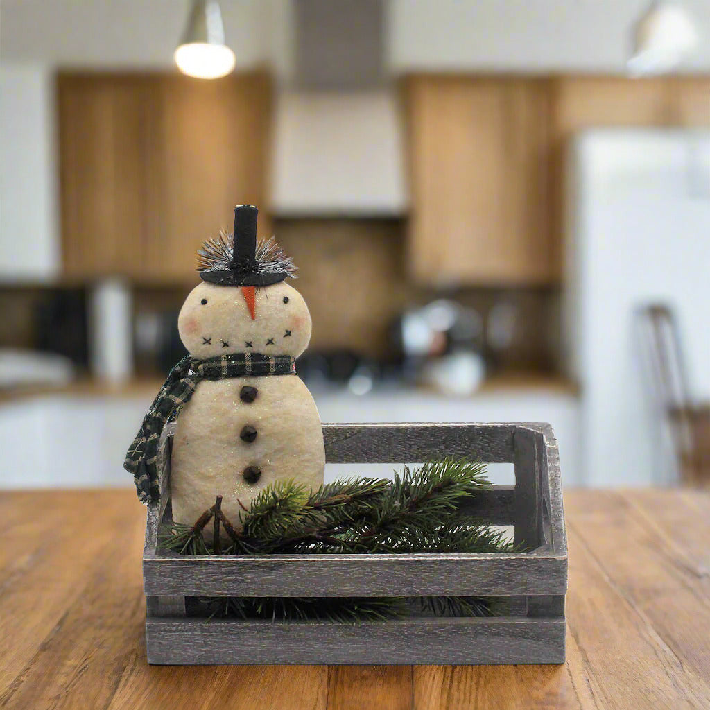 Decorative snowman figurine with a black top hat and scarf, placed in a wooden crate with greenery