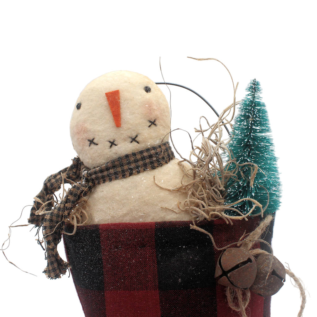 Decorative snowman figurine with a plaid scarf, carrot nose, and small tree on a white background.