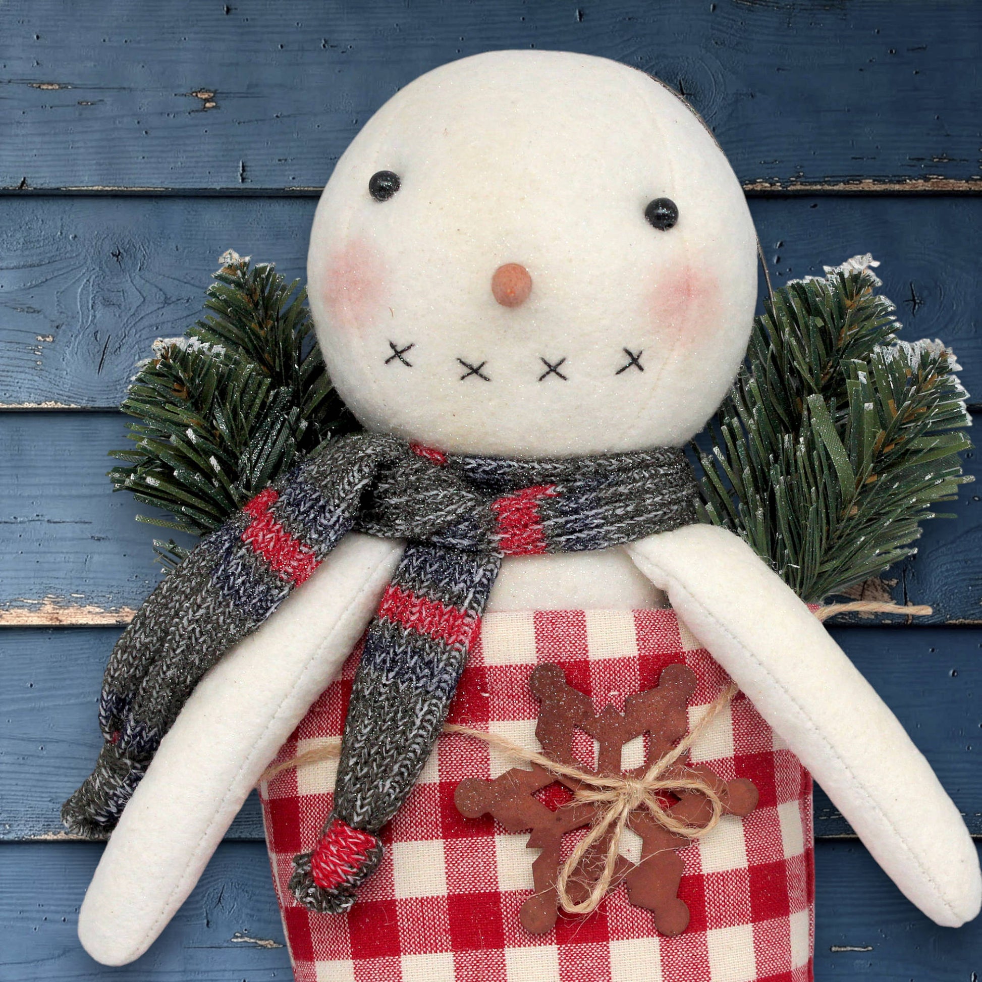 Textile snowman with a red and white checkered scarf and plaid pants against a blue wooden background
