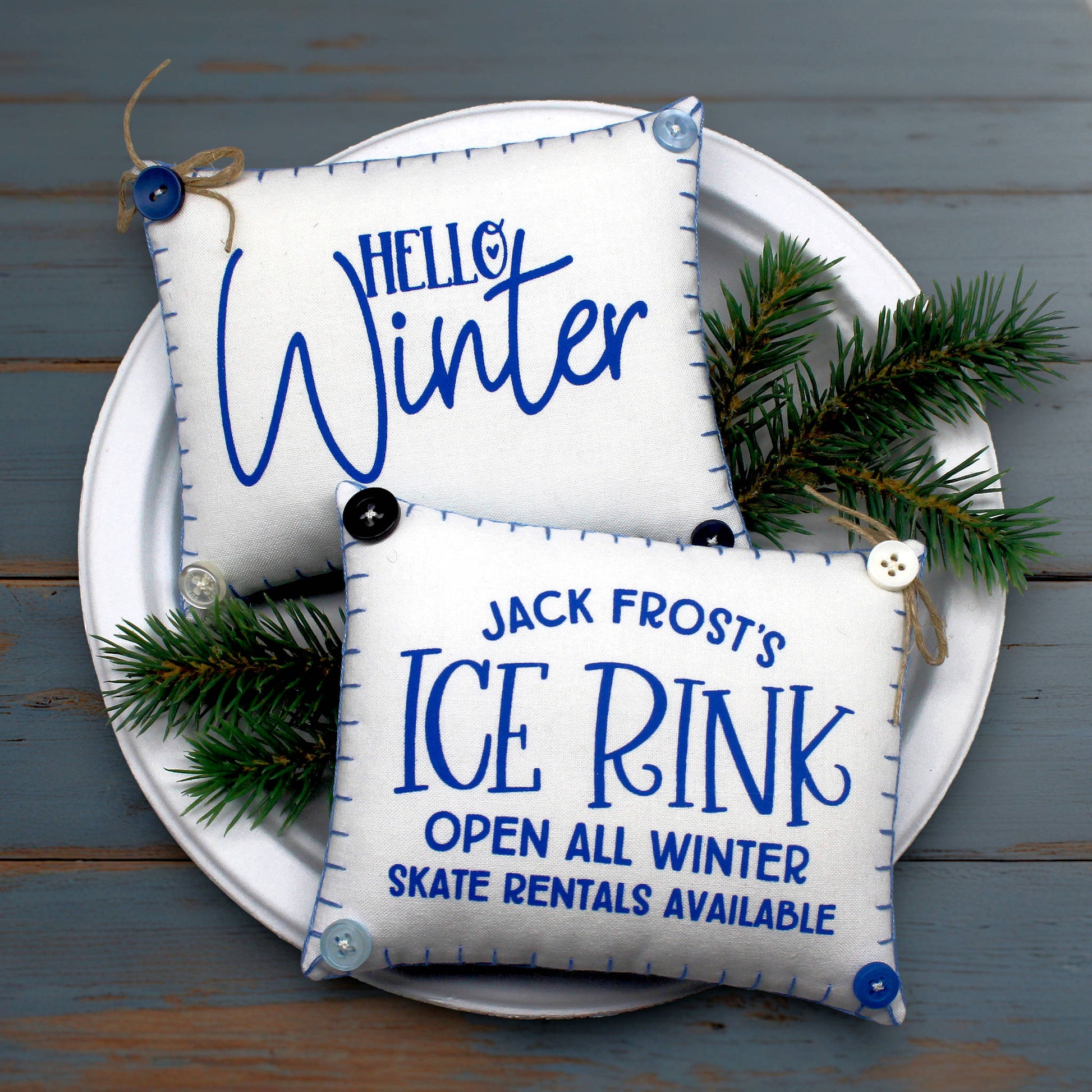 Two decorative mini pillow bowl fillers with winter-themed text on a white plate with greenery against a wooden background.