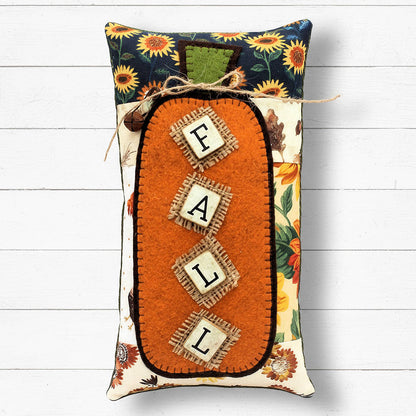 Decorative pillow with 'FALL' letters and autumn-themed design on a white wooden background