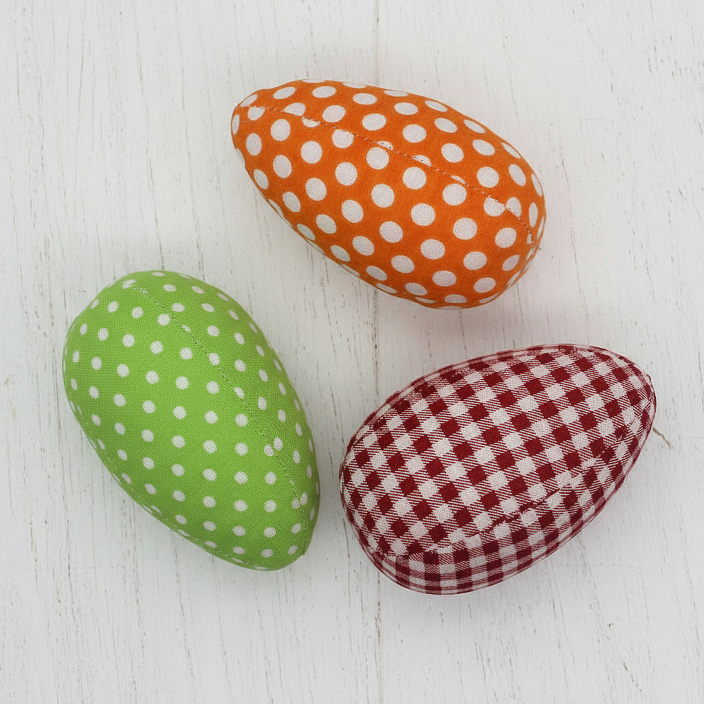 Three fabric Easter eggs in orange with white polka dots, green with white polka dots, and red with white checkered pattern on a white background.