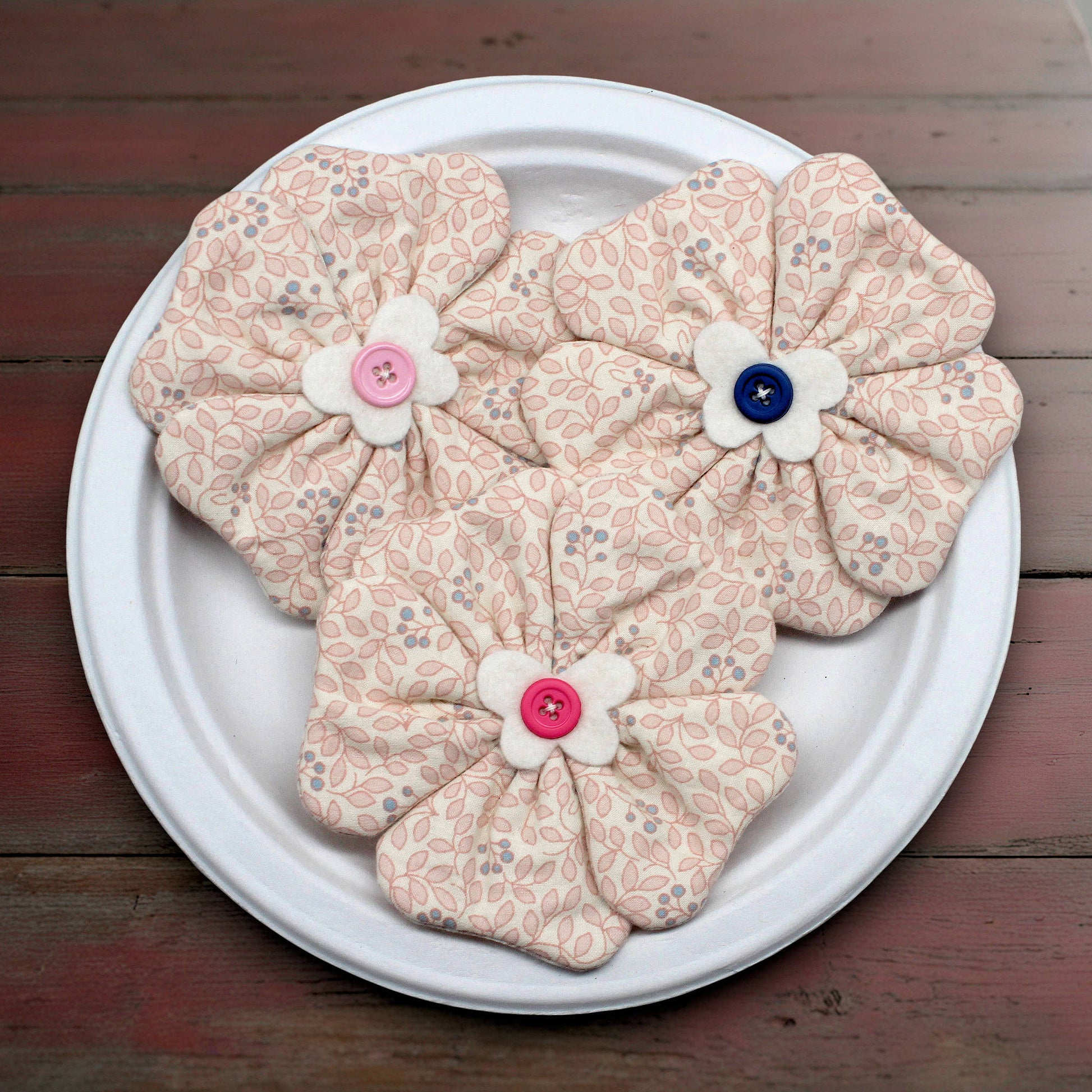 Floral-patterned fabric flowers with buttons on a white plate against a wooden background