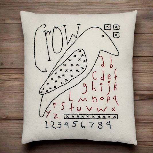 Decorative pillow with embroidered crow sampler design on a wooden surface