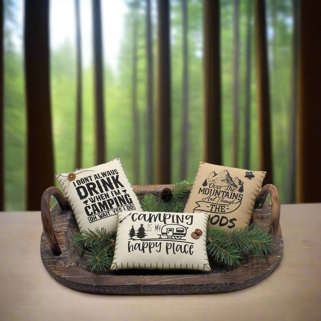 Three decorative pillows with camping-themed text on a wooden tray with pine branches.