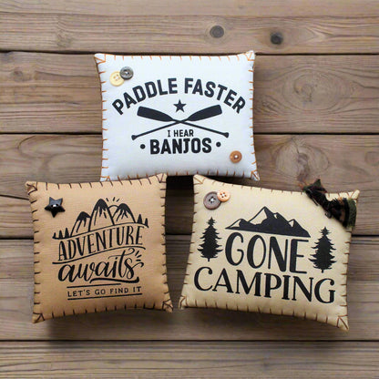 Three decorative pillows with camping-themed text on a wooden surface