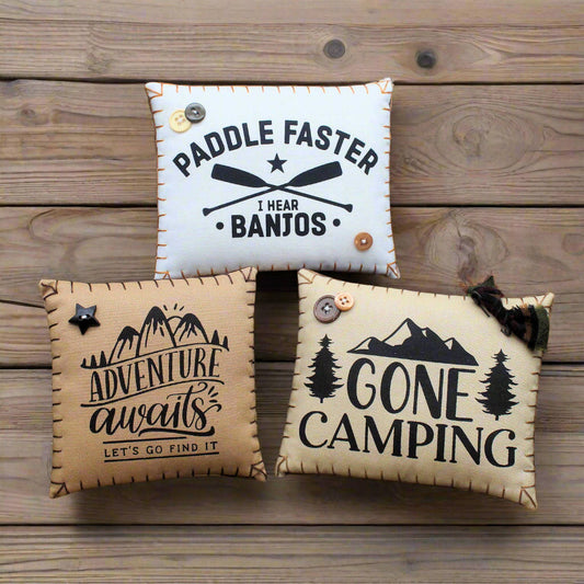 Three decorative pillows with camping-themed text on a wooden surface