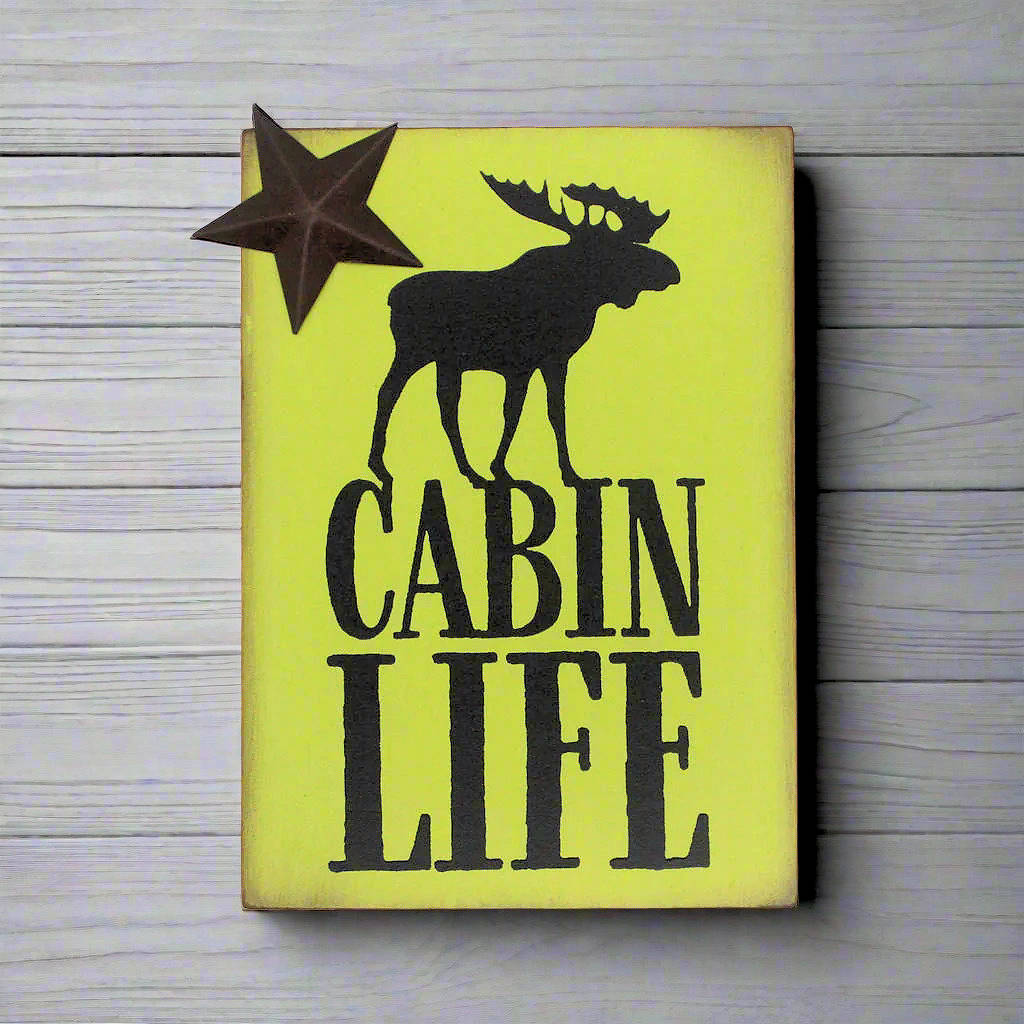 Wooden sign with a moose silhouette and 'Cabin Life' text on a yellow green background, placed on a wooden surface.
