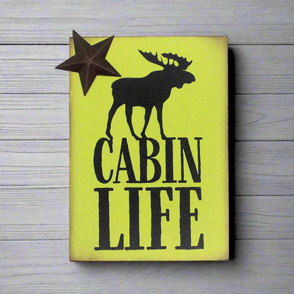 Wooden sign with a moose silhouette and 'Cabin Life' text on a yellow green background, placed on a wooden surface.