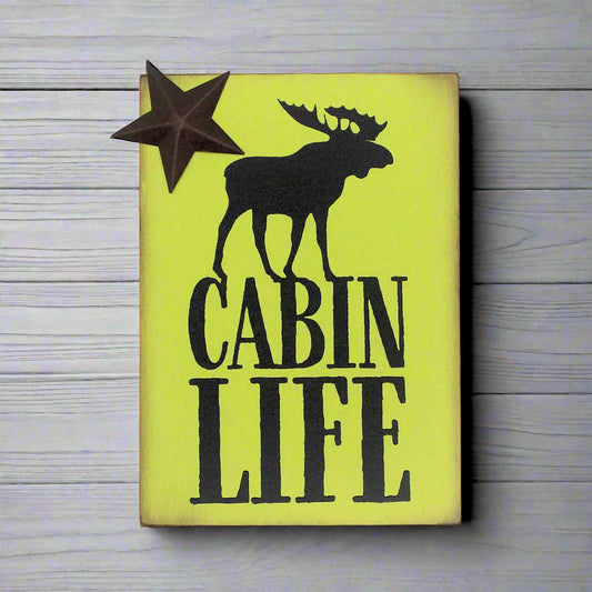 Wooden sign with a moose silhouette and 'Cabin Life' text on a yellow green background, placed on a wooden surface.