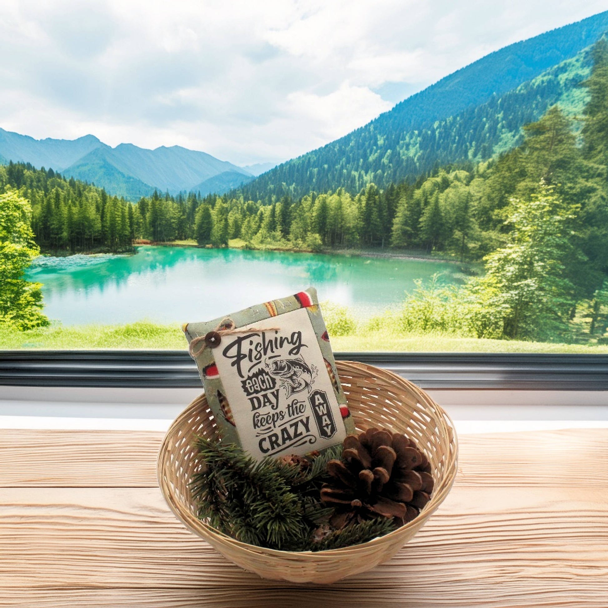 Decorative pillow tuck with text in a woven basket on a windowsill with a scenic view of mountains and lake.