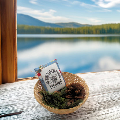 A basket with a fishing-themed pillow tuck, pine and pine cones on a windowsill looking out onto a mountain lake