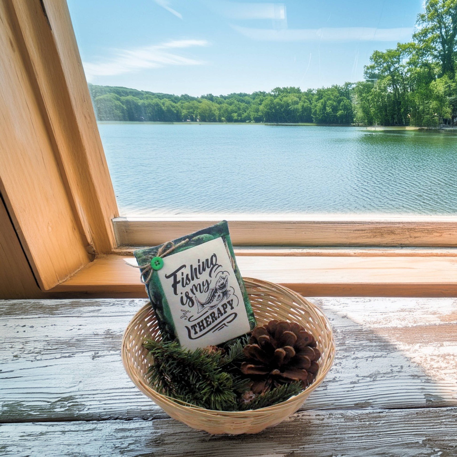 Decorative basket with a sign and pinecones on a windowsill with a lake view.