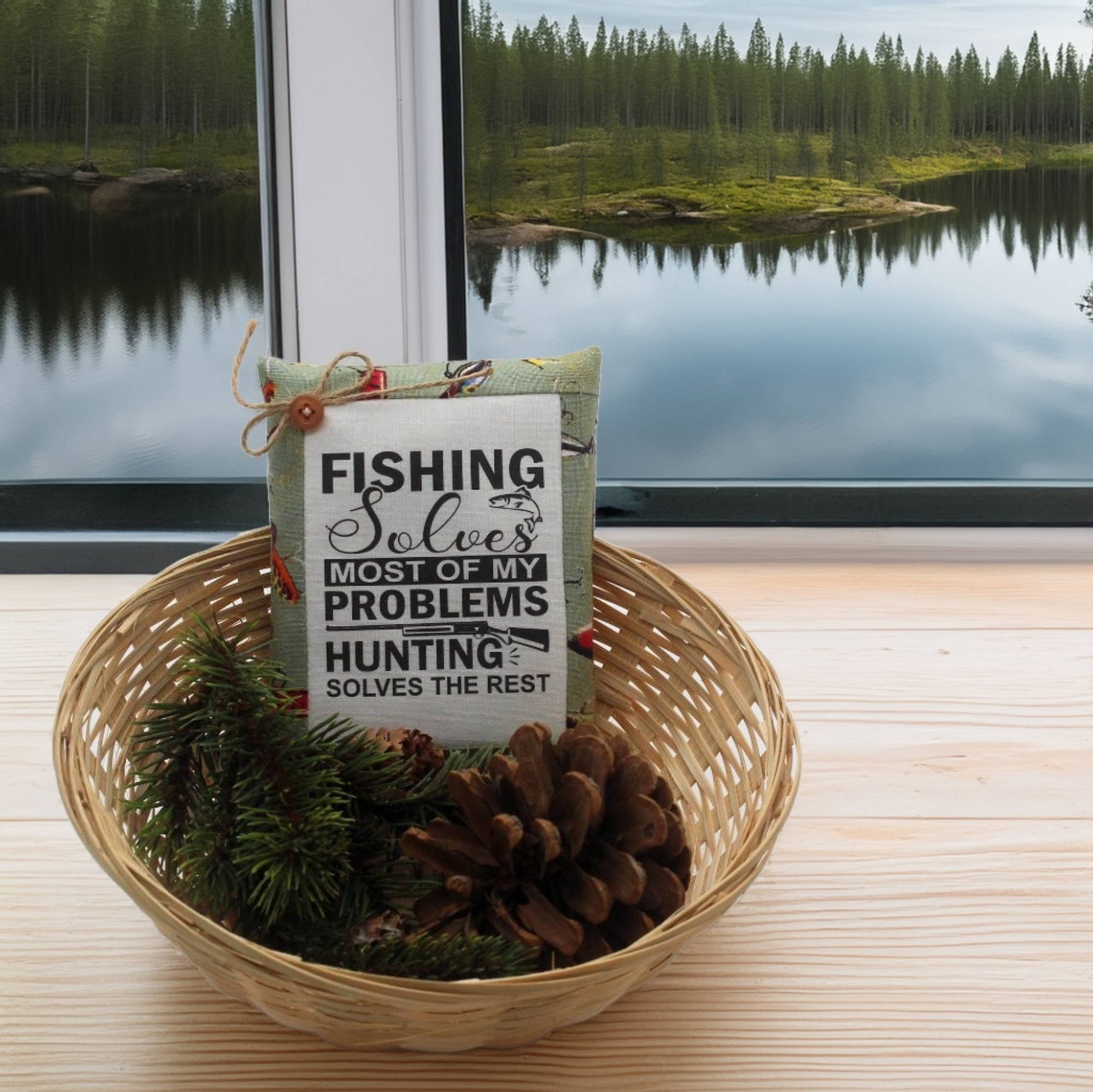 Decorative pillow with text in a woven basket with pine cones and branches, set against a scenic window view of a lake and forest.