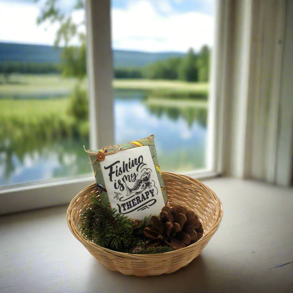 Decorative sign with 'Fishing is my therapy' text in a woven basket with moss and pinecones, set against a window view of a lake and forest.