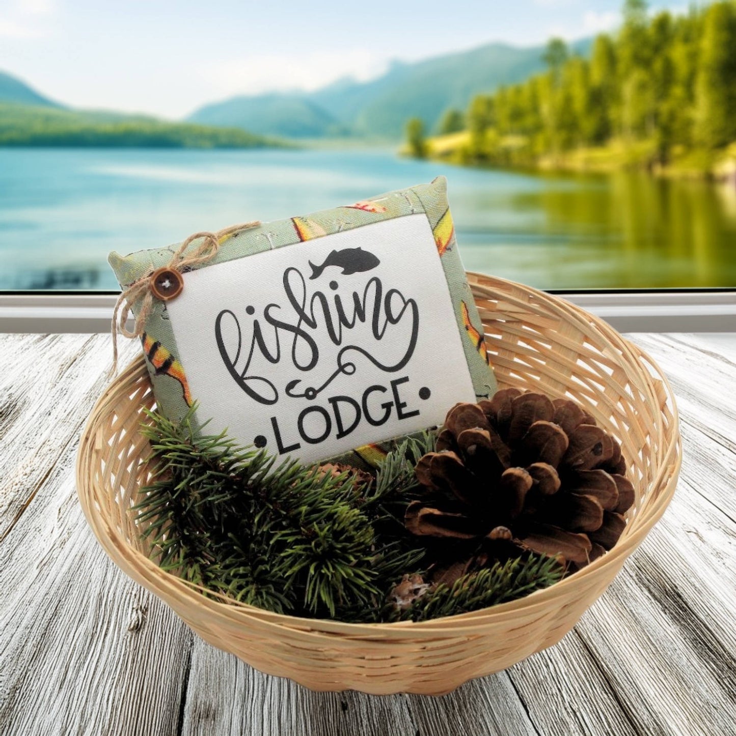 Decorative basket with 'Fishing Lodge' sign, pinecones, and greenery against a scenic lake and mountain background.