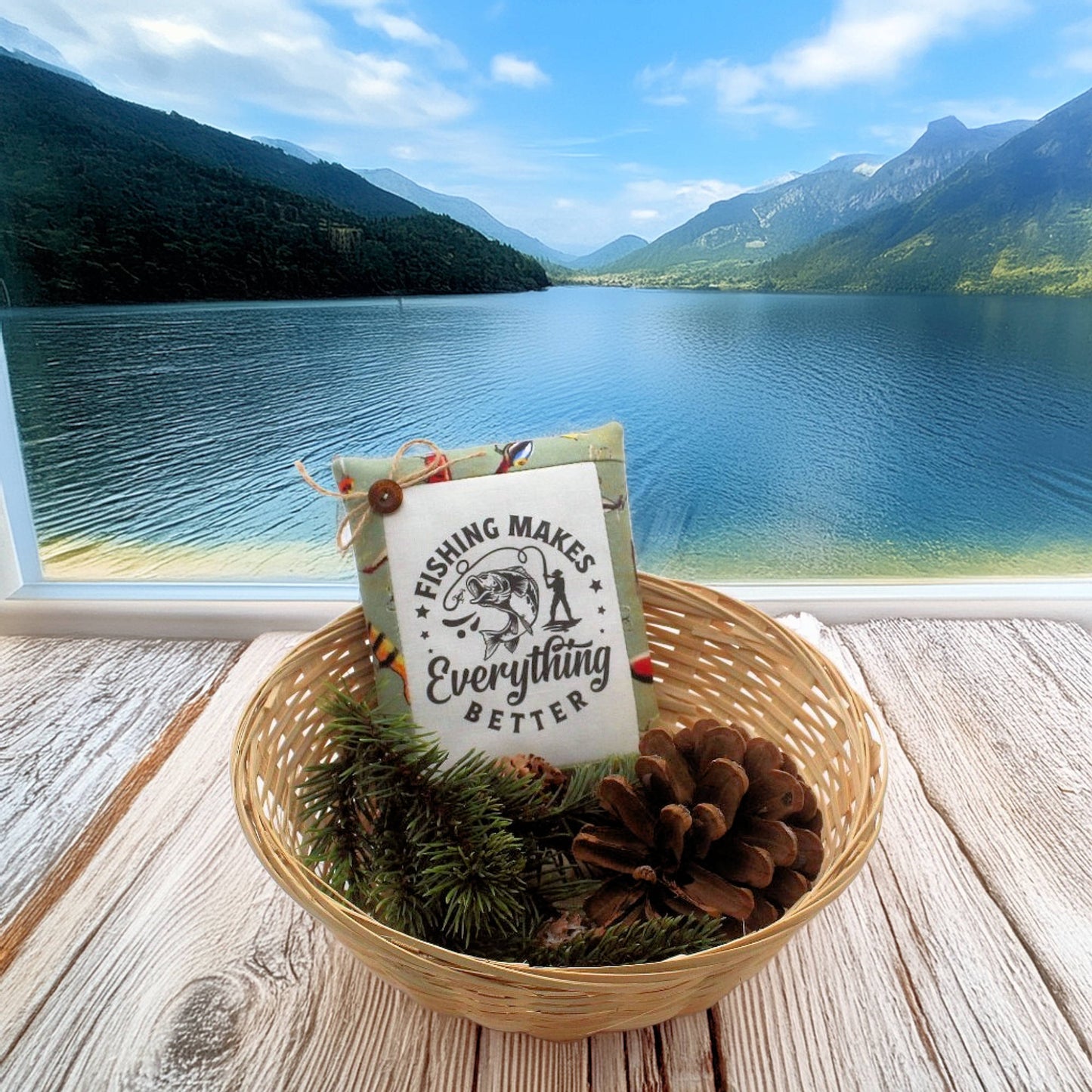 Decorative pillow with 'Fishing Makes Everything Better' text in a woven basket by a lake with mountains.