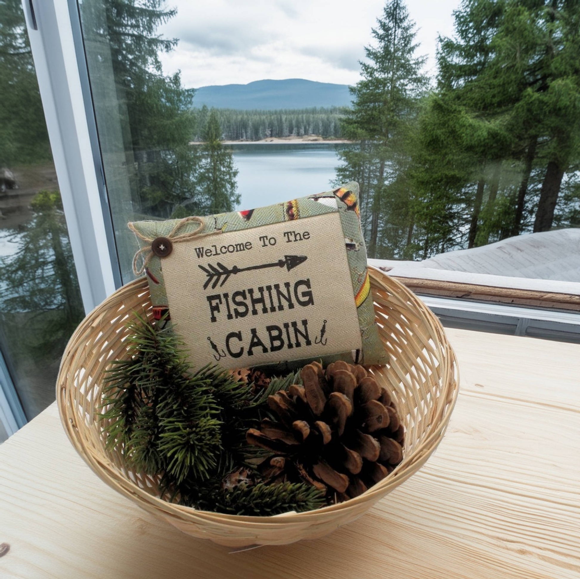 Decorative basket with a 'Welcome to the Fishing Cabin' sign, pinecones, and evergreen branches on a windowsill with a view of a lake and forest.