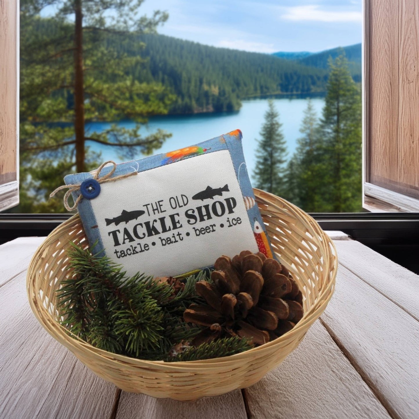 Decorative sign with 'The Old Tackle Shop' text in a basket with pine branches and pinecones, set against a scenic window view of a lake and forest.