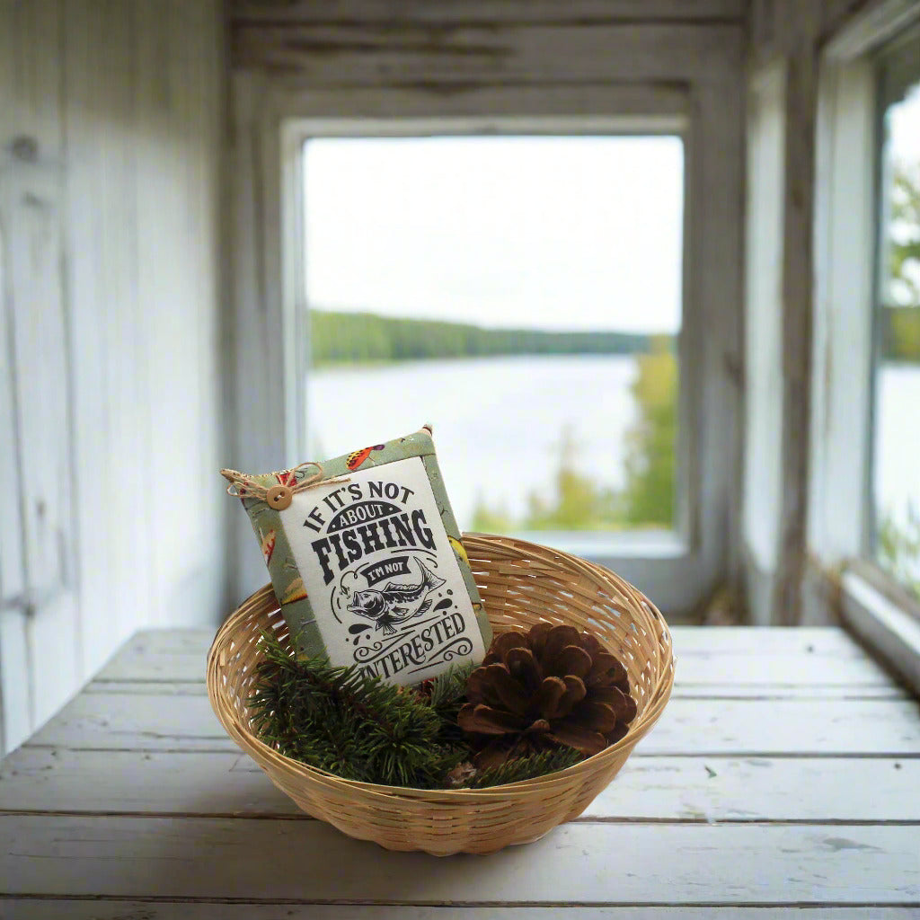 Decorative basket with a sign and pinecones on a wooden table by a window with a view of water.