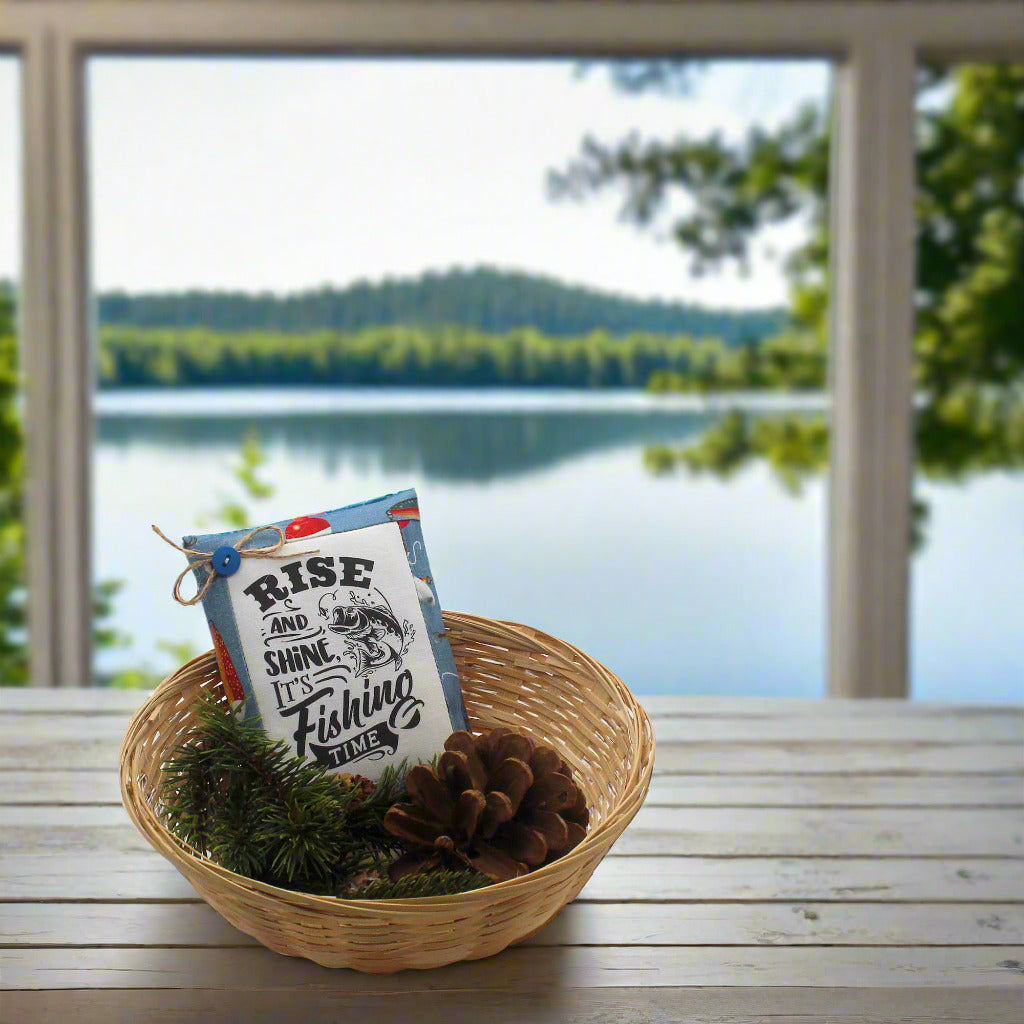 Basket with a 'Rise and Shine, It's Fishing Time' sign on a wooden table with a lake view.