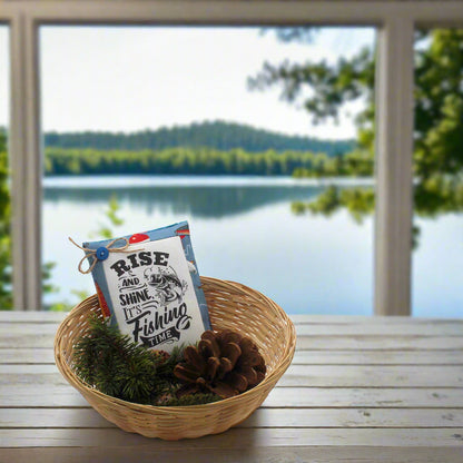 Basket with a 'Rise and Shine, It's Fishing Time' sign on a wooden table with a lake view.