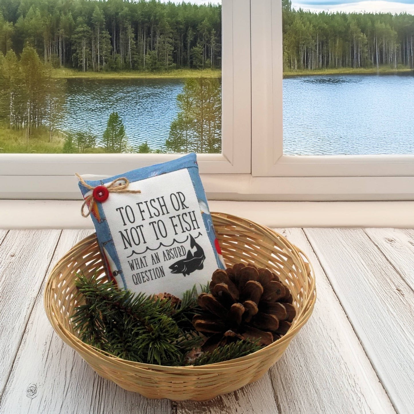 Decorative pillow with fishing theme in a basket on a wooden floor with a window view of a lake and forest.