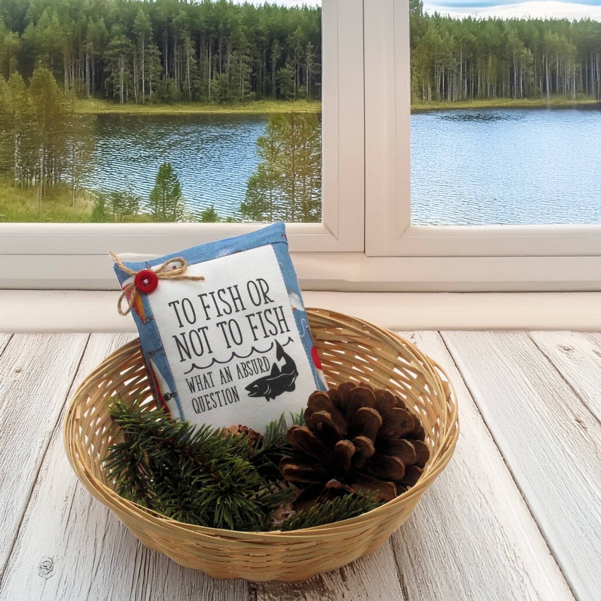 Decorative pillow with fishing theme in a basket on a wooden floor with a window view of a lake and forest.