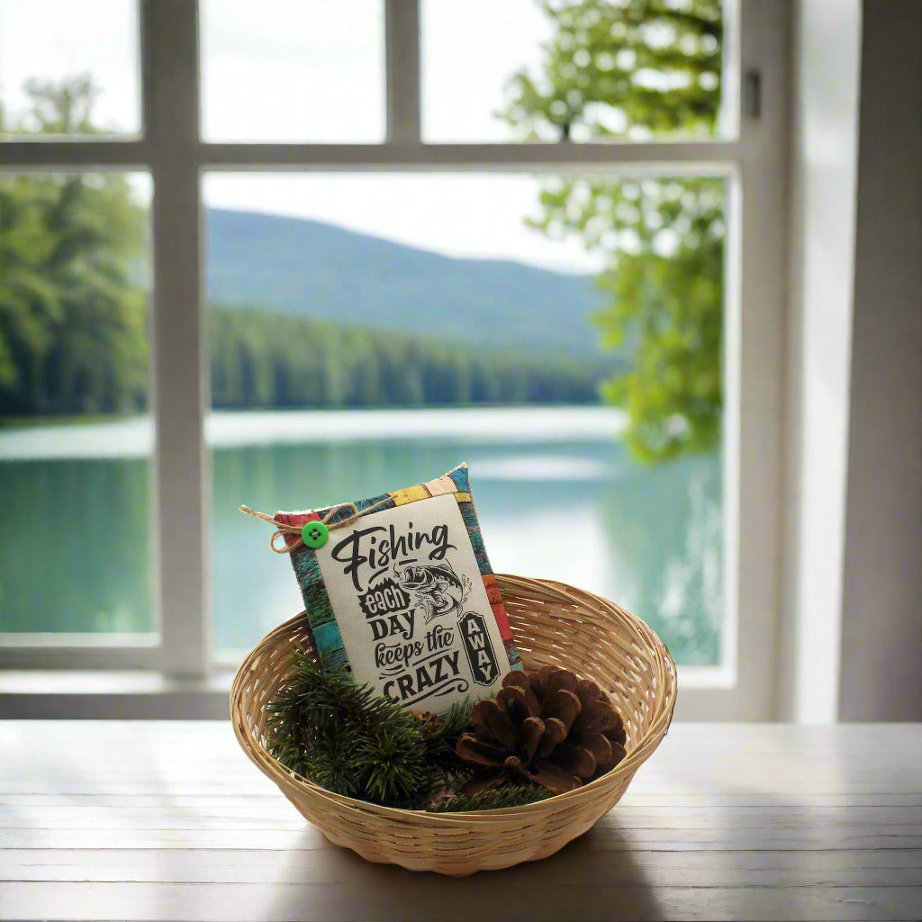 Decorative pillow with text and graphics in a woven basket on a table with a scenic view of a lake through a window.