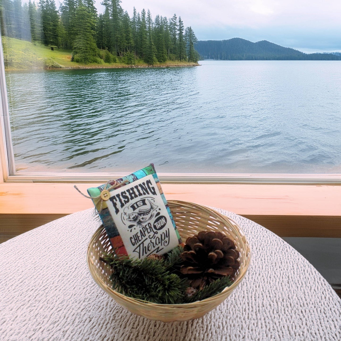 Decorative basket with a pillow and pinecones on a table by a window overlooking a lake and forest.