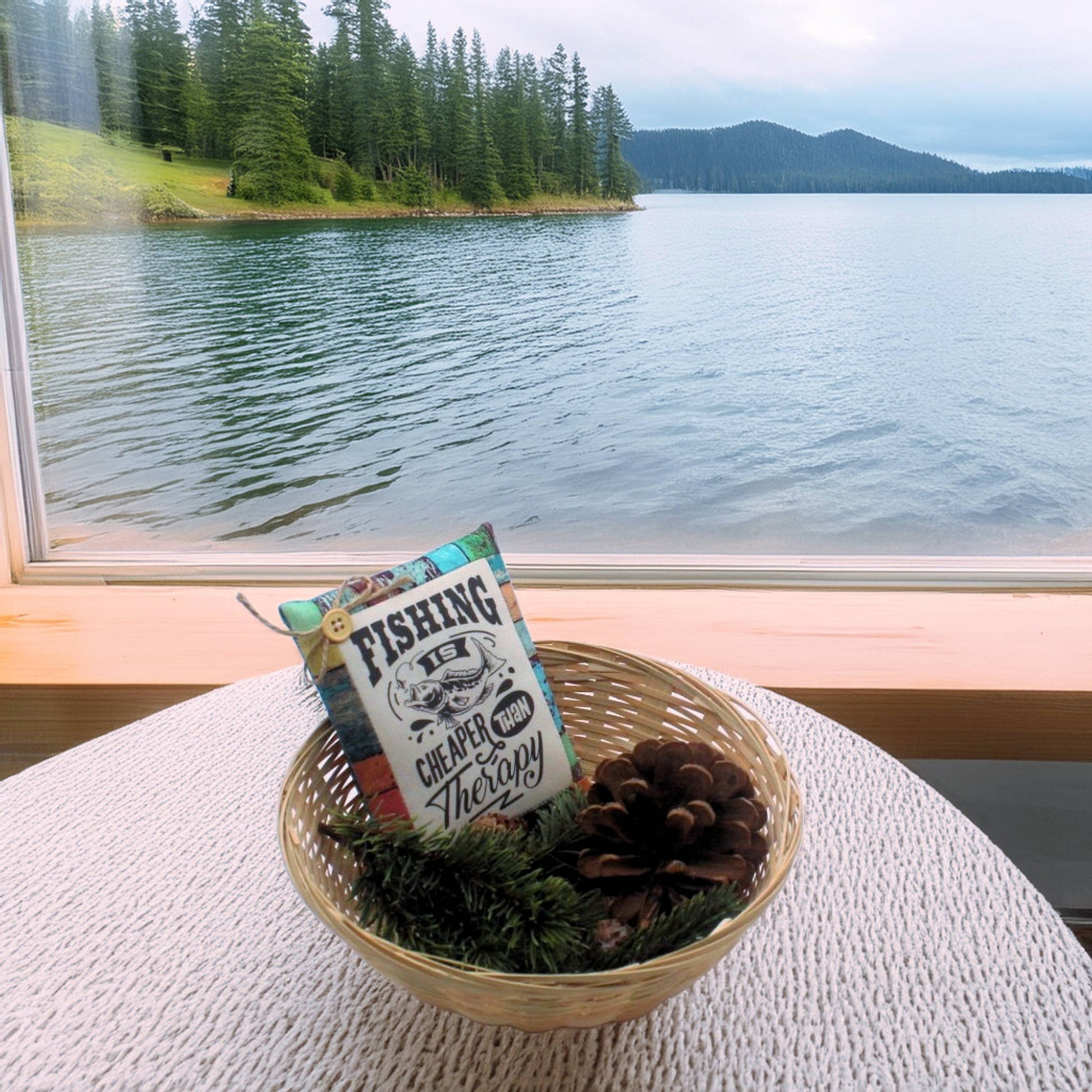 Decorative basket with a pillow and pinecones on a table by a window overlooking a lake and forest.