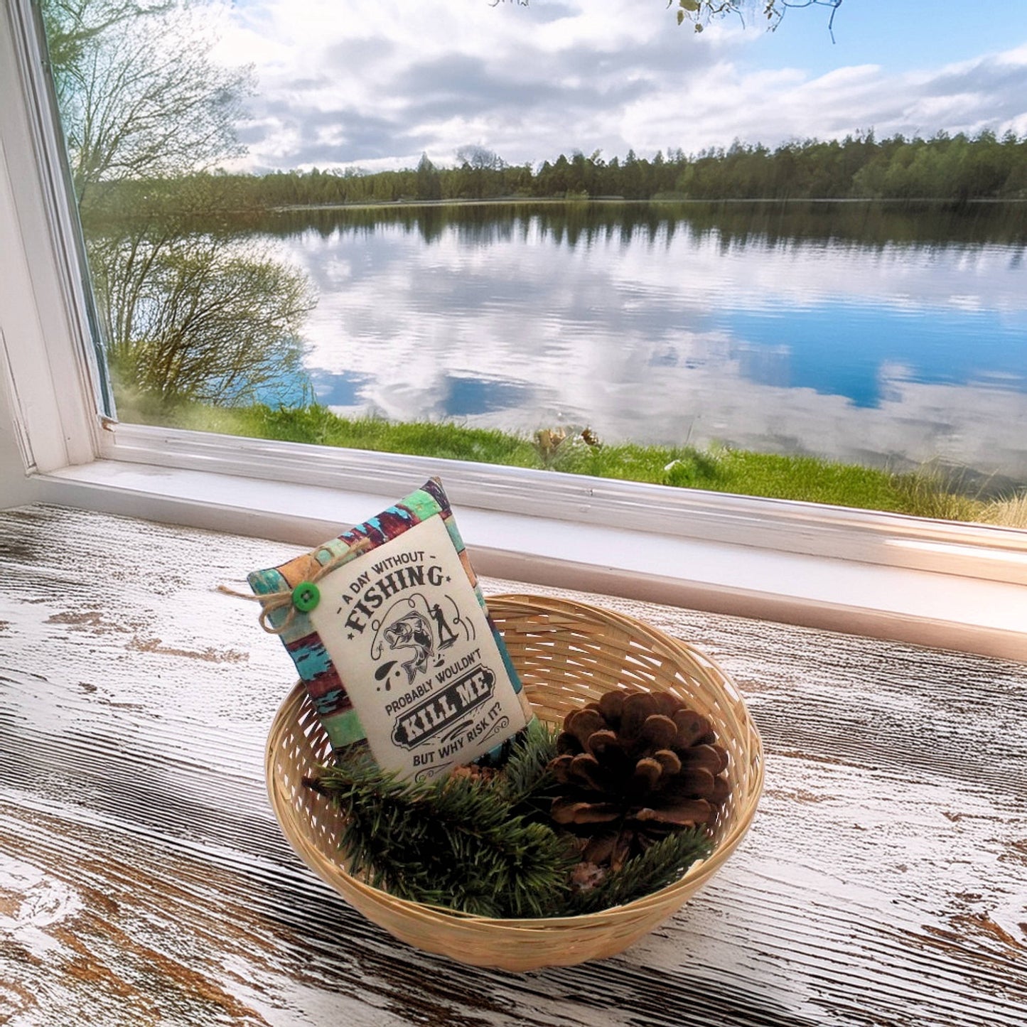 Decorative pillow with fishing design in a woven basket by a window with a view of a lake.