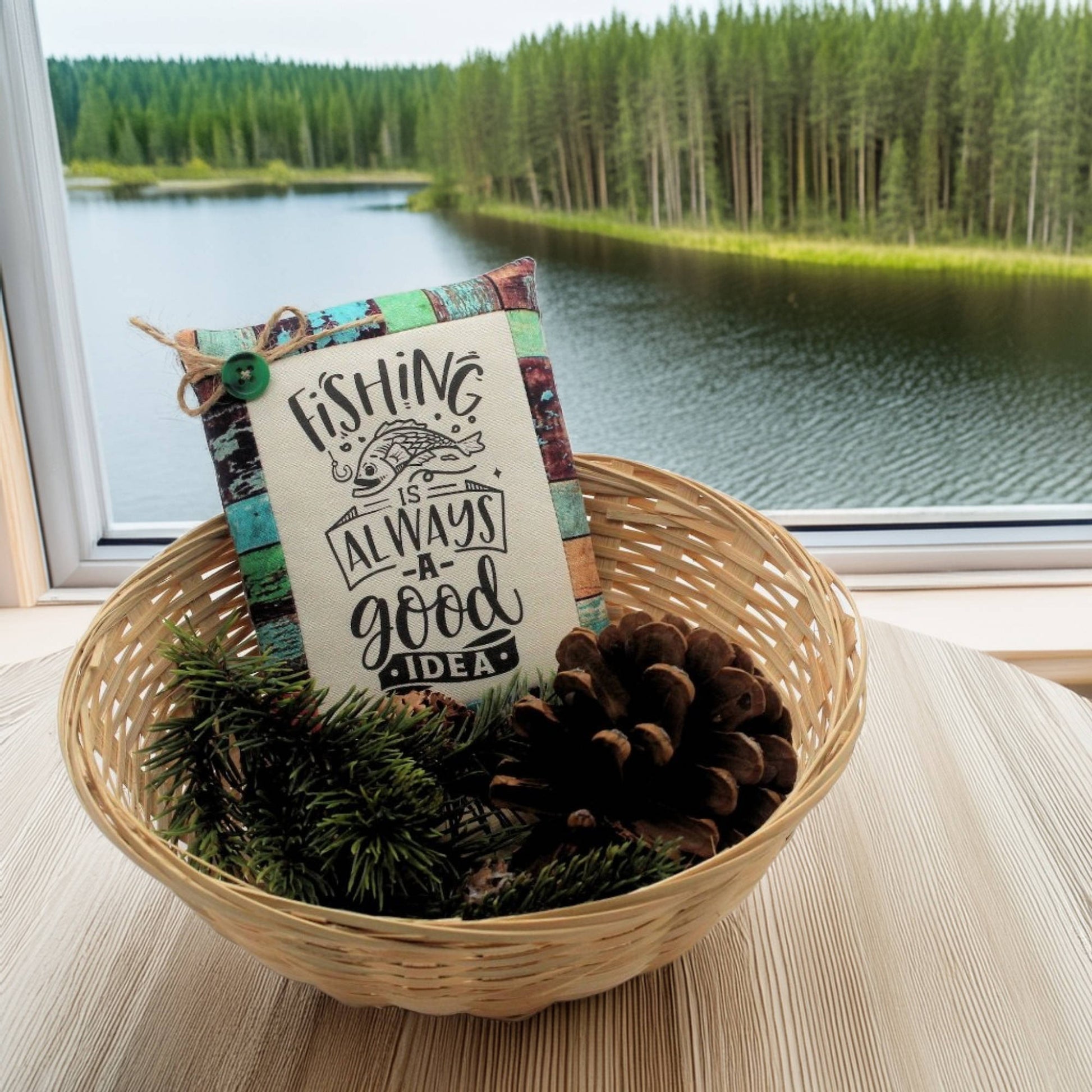 Decorative sign with 'Fishing is always a good idea' text in a woven basket with pinecones and greenery, set against a scenic lake and forest background.