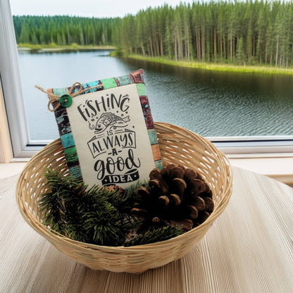 Decorative sign with 'Fishing is always a good idea' text in a woven basket with pinecones and greenery, set against a scenic lake and forest background.