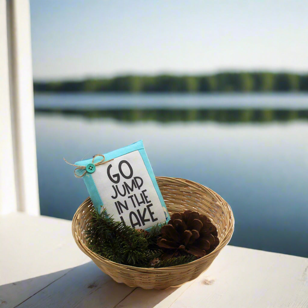 Decorative sign with 'Go jump in the lake' text in a woven basket by a lakeside.
