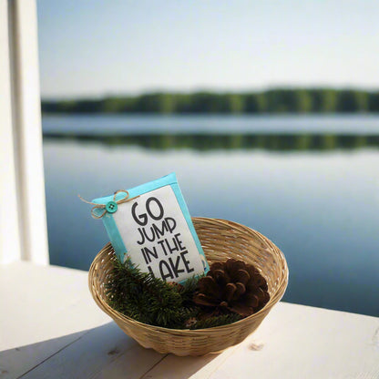 Decorative sign with 'Go jump in the lake' text in a woven basket by a lakeside.