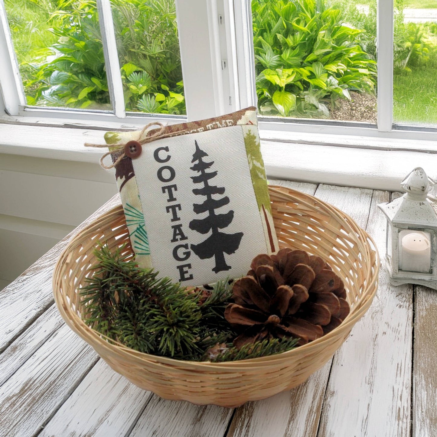 Decorative basket with a 'Cottage' sign, pinecones, and greenery on a wooden surface.