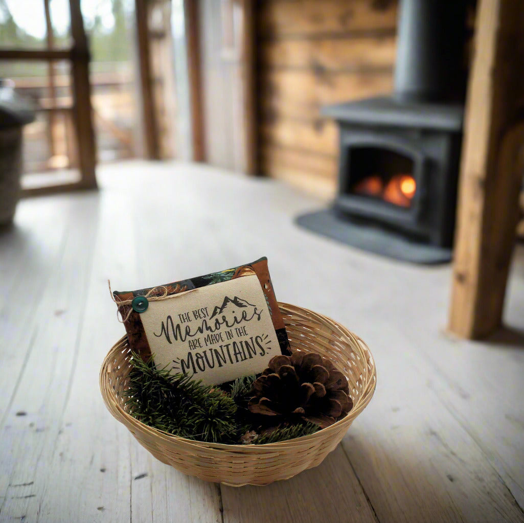 Decorative basket with a sign and pine cone on a wooden floor in front of a fireplace.