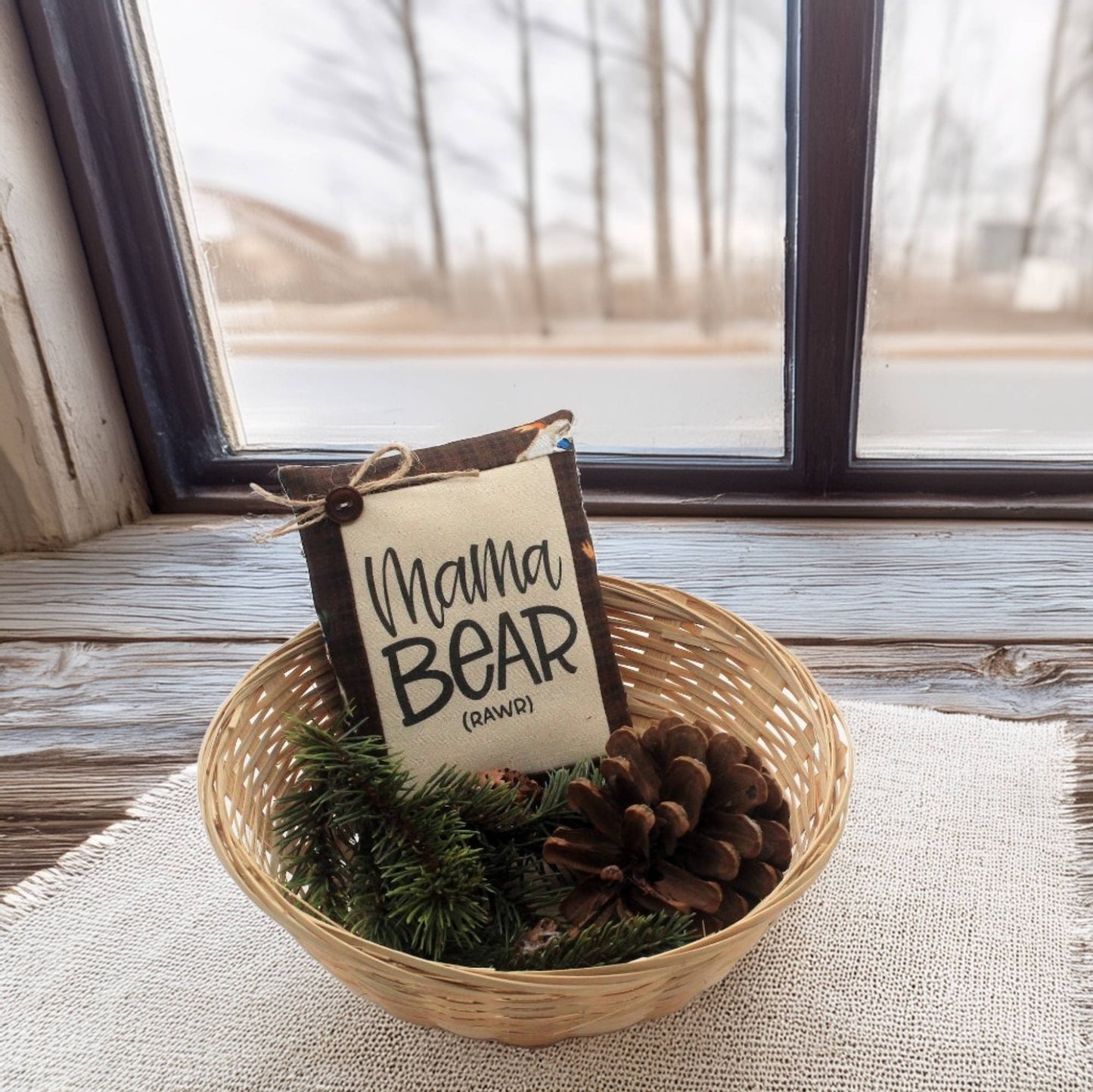 Decorative item with 'Mama Bear' sign in a basket on a windowsill.
