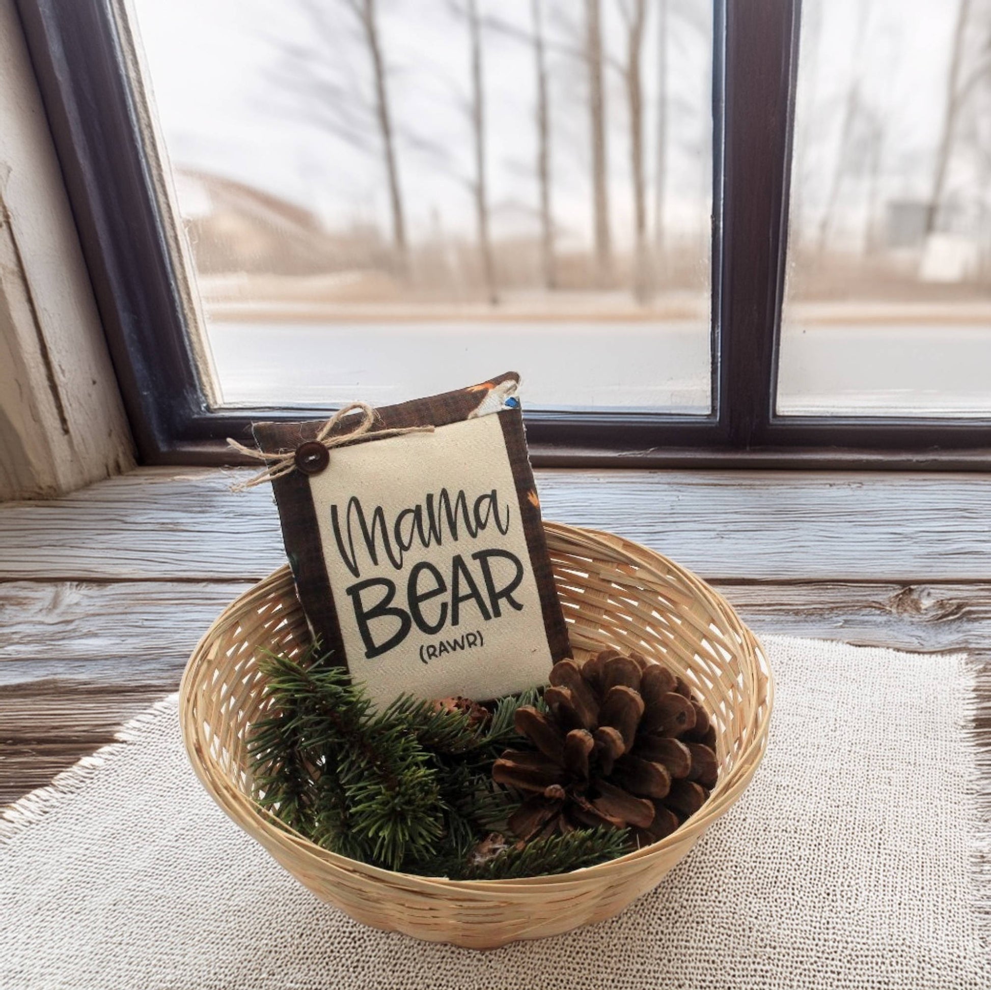 Decorative item with 'Mama Bear' sign in a basket on a windowsill.