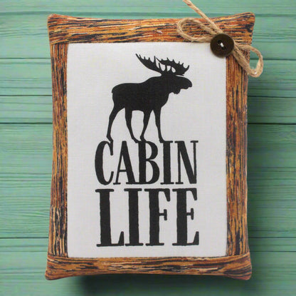 Decorative mini pillow sign with moose silhouette and 'Cabin Life' text on a wooden frame against a green background.