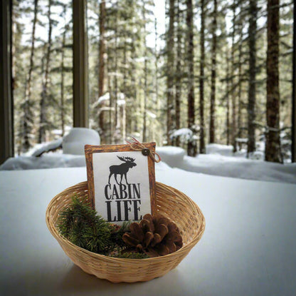Basket with 'Cabin Life' sign and moose illustration in a snowy forest setting