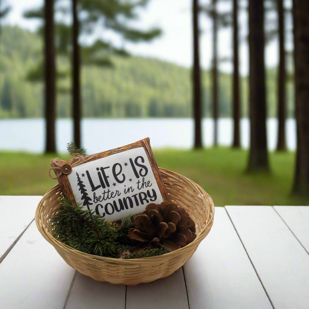Decorative sign with 'Life is better in the country' text in a basket with greenery on a wooden table by a lake.